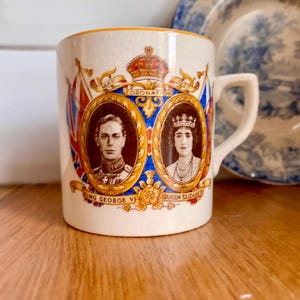 Może przedstawiać: Zabytkowy ceramiczny kubek z portretami króla Jerzego VI i królowej Elżbiety, ze słowem „CORONATION” i królewską koroną. Kubek ma kremowe tło ze złotymi, czerwonymi i niebieskimi akcentami. Ozdobny talerz znajduje się w tle.