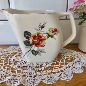 Puede incluir: Una jarra de cerámica blanca con un borde dorado y un diseño floral con rosas rojas, amarillas y verdes. La jarra tiene un asa y un pico.