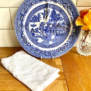 Op de afbeelding: Een blauw-wit keramisch bord met een gedetailleerd landschapsontwerp, tentoongesteld op een houten oppervlak. Een witte, gevouwen linnen servet ligt op de voorgrond. Een vaas met gele rozen staat op de achtergrond.