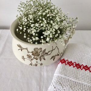 Könnte beinhalten: Eine weiße Keramikschale mit Blumenmuster und einem Strauß weißer Baby's Breath-Blumen. Die Schale hat ein braunes Blumenmuster und steht auf einer weißen Tischdecke.
