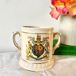 Op de afbeelding: Een witte keramische mok met gouden rand en een kleurrijk koninklijk wapen met een leeuw, een eenhoorn en een schild. De mok herdenkt de kroning van koningin Elizabeth II op 2 juni 1953.
