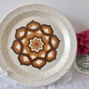 May include: A white ceramic dinner plate with a brown, orange, and white geometric floral design. The plate has a speckled texture.