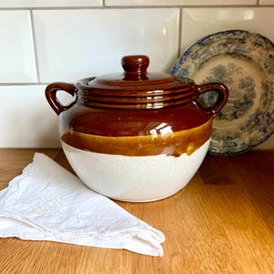 May include: A round ceramic casserole dish with a lid, featuring a two-tone design with a glossy dark brown glazed upper section and a speckled cream-coloured lower base. The pot includes two side handles and a small knob on the lid.