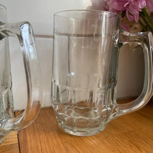 Op de afbeelding: Twee heldere glazen bierpullen met handvatten. De pullen hebben een rechthoekig ontwerp op de onderste helft. De pullen staan op een houten oppervlak. Een vaas met roze bloemen staat op de achtergrond.