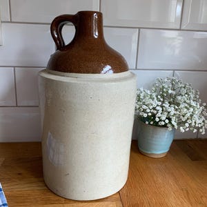 May include: A vintage stoneware jug with a brown top and handle, and a cream-colored base. The jug is cylindrical and sits on a wooden surface. A small blue pot with white flowers is to the right.