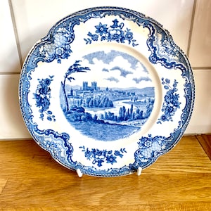 Pode incluir: Um prato de cerâmica azul e branco com uma borda floral e uma cena de uma cidade com um rio e árvores no centro. O prato está sobre uma superfície de madeira.