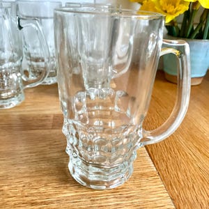 May include: Clear glass beer mugs with textured bases and handles. Several mugs are arranged on a wooden surface. Yellow flowers in a blue pot are in the background.