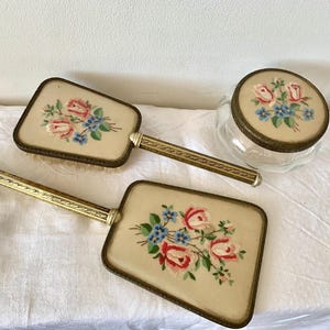May include: A vintage vanity set featuring two hand mirrors and a small jar. The set has a cream-colored background with embroidered red, pink, and blue floral designs. The handles and jar rim are gold-toned.