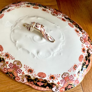Op de afbeelding: Een witte keramische serveerschaal met een rood en gouden bloemmotief rond de rand. De schaal heeft een verhoogd, geëmbost midden met een rood en gouden handvat.