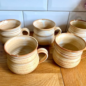 May include: Five handmade ceramic mugs with a rustic, earthy aesthetic. The mugs feature a warm, beige colour with brown, horizontal stripes and a curved handle. The mugs are arranged on a wooden surface, suggesting a cosy, homey feel.