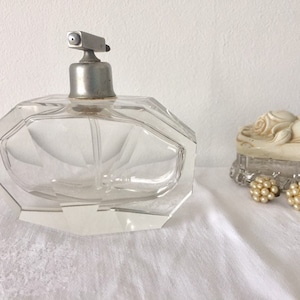 Könnte beinhalten: Eine durchsichtige Glas-Parfümflasche mit einem silbernen Zerstäuber und einem facettierten Design. Die Flasche steht auf einer weißen Oberfläche.