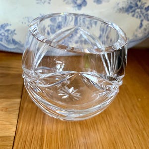 Puede incluir: Un jarrón de vidrio transparente con un diseño de cristal tallado. El jarrón es redondo y tiene una abertura ancha.