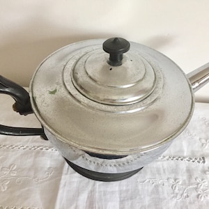 Könnte beinhalten: Eine antike silberne und schwarze elektrische Kaffeekanne mit schwarzem Griff und silbernem Deckel. Die Kaffeekanne steht auf einer weißen Spitzentischdecke.