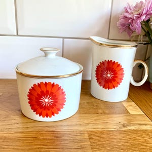 May include: A white ceramic sugar bowl with a lid and a creamer with a handle. Both pieces have a red and orange floral design and gold trim. The set is on a wooden surface.