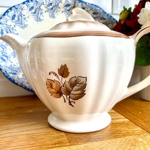 Puede incluir: Una tetera de cerámica blanca con tapa y asa marrones. La tetera tiene un diseño floral marrón con hojas y un borde marrón alrededor de la tapa y el pico.