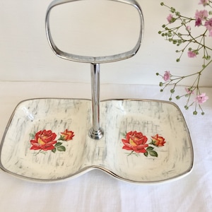 May include: Two-tiered serving tray with a silver handle. The tray has two sections with a cream-colored background and a gray, textured pattern. Each section features a red rose design with green leaves.