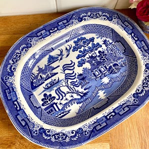 May include: An oval ceramic serving platter featuring a blue and white willow pattern. The design depicts a traditional landscape with pagodas, a bridge, a boat, and stylized trees. The wide rim has a geometric border pattern in blue and white.