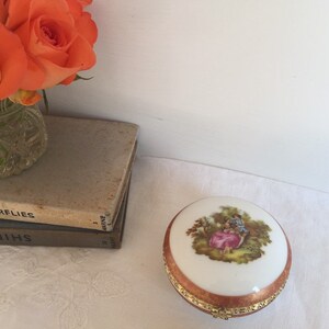 Puede incluir: Una caja de porcelana blanca con borde dorado y una escena pintada de una pareja en un jardín. La caja está sobre una superficie blanca.