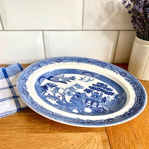 Puede incluir: Plato ovalado de cerámica azul y blanco con un diseño de patrón Willow. El plato está sobre una superficie de madera junto a un paño a cuadros azul y blanco y una maceta de cerámica blanca con lavanda.