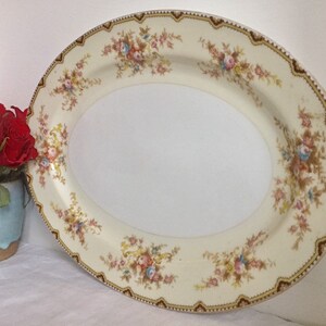 May include: A large oval serving platter with a cream-colored background and a floral pattern in shades of pink, yellow, and blue. The border of the platter is decorated with a gold and brown trim.