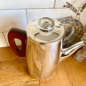 Può includere: Una caffettiera argentata lucida con coperchio in vetro e manico in legno scuro. La caffettiera ha un beccuccio ed è appoggiata su una superficie di legno. Un rametto di lavanda e un piatto decorativo sono sullo sfondo.