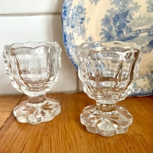May include: Two clear, vintage glass egg cups with scalloped rims and starburst bases. A blue and white decorative plate with a landscape design is in the background. The egg cups are on a wooden surface.