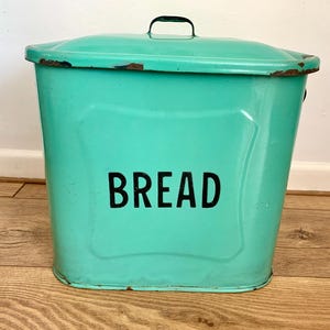 May include: A vintage-style turquoise metal bread bin with a matching lid and a small black handle. The front of the container features the word BREAD printed in bold black capital letters. The edges show signs of wear with visible rust spots.