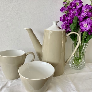 以下が含まれることがあります： ベージュのセラミック製のティーポット、クリーマー、シュガーボウルセット。ティーポットは白い蓋と注ぎ口が付いています。クリーマーとシュガーボウルはどちらも丸く、シンプルなデザインです。