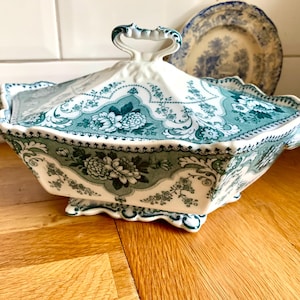 Puede incluir: Una fuente de cerámica blanca y verde azulado con tapa, con diseños florales y de volutas. La fuente tiene forma rectangular con borde festoneado y asa. Un plato a juego se ve en el fondo.