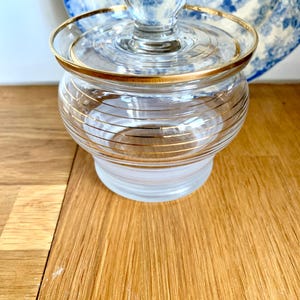 Könnte beinhalten: Ein klares Glasgefäß mit Deckel, verziert mit goldenen Zierleisten. Das Gefäß hat horizontale Goldstreifen und eine abgerundete Form. Der Deckel hat einen kleinen Knopf. Im Hintergrund befindet sich ein blau-weißes Zierteller.