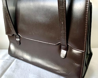May include: A dark brown leather handbag with a glossy finish. The bag has a rectangular shape with a flap closure and two short handles. The bag has a structured design and appears to be vintage.