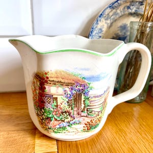 May include: White ceramic pitcher with a scalloped rim and a green border. The pitcher features a colorful illustration of a cottage with a thatched roof and blooming flowers. It has a curved handle.