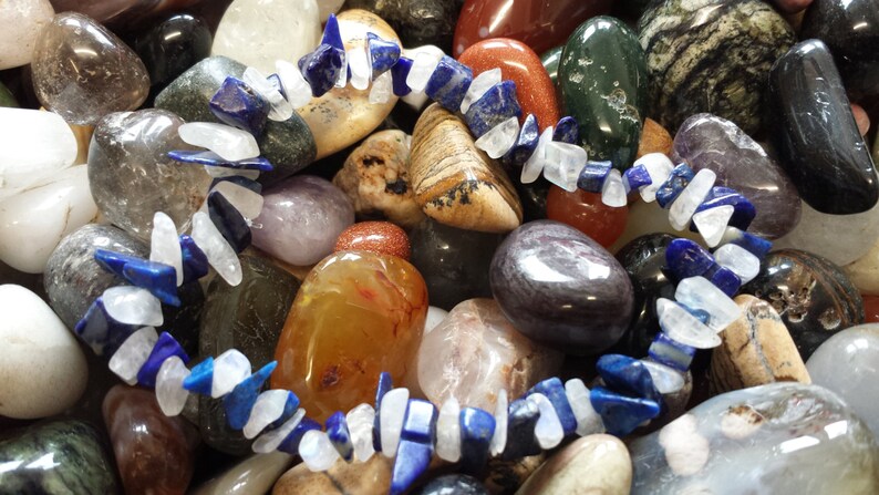 May include: A bracelet made of white and blue gemstone beads. The bracelet is laid out on a bed of various colorful stones.