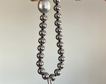 May include: A necklace featuring a strand of polished metallic silver beads strung on a dark cord, accented by a single large, smooth, light gray pearl positioned asymmetrically, and finished with a small silver-toned metal clasp at the end.