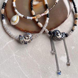 Op de afbeelding: Verschillende kettingen met kralen en details van schelpen en keramische hangers. De kettingen hebben een verscheidenheid aan kralen in wit, zwart, bruin en roze. Eén ketting heeft een schelpenhanger, andere hebben keramische hangers.