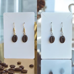 May include: Two pairs of coffee bean dangle earrings. One pair has gold hooks and the other has silver hooks. The coffee beans are brown and the earrings are displayed on white cards.