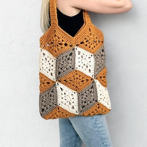 Op de afbeelding: Een handgemaakte gehaakte tote bag met een geometrisch kubuspatroon. De tas heeft een combinatie van mosterdgele, crème en grijze vierkanten. De tas heeft een mosterdgele band en wordt over de schouder gedragen.