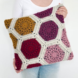 Op de afbeelding: Een handgemaakt gehaakt kussen met een geometrisch patroon van zeshoeken in mosterdgeel, bordeauxrood, roze en crème. Het kussen wordt vastgehouden door een persoon die een wit shirt met lange mouwen en een blauwe spijkerbroek draagt.