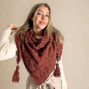 May include: A rust-colored, crocheted shawl with a triangular shape and tassel accents. The shawl is made of a textured yarn and is draped around the neck. The tassels hang from the points of the shawl.