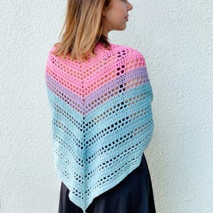 Op de afbeelding: Een handgemaakte gehaakte sjaal in een verloop van roze, lavendel en blauw. Het open weefselontwerp heeft een chevronpatroon. Een bijpassende blauwe kwast hangt aan de onderkant van de sjaal. De sjaal wordt over een zwarte jurk gedragen.