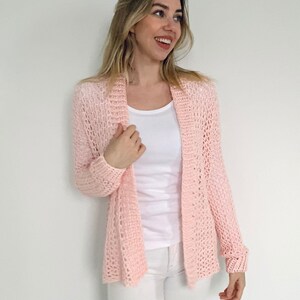 May include: A light pink, open-front crochet cardigan featuring long sleeves and a textured, airy stitch pattern. The garment is worn over a plain white scoop-neck top and white pants, showcasing a lightweight, breathable knit construction.