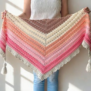 Op de afbeelding: Een handgemaakte, kleurrijke gehaakte sjaal met een verloop van roze, oranje en beige tinten. De sjaal heeft een delicate kanten rand en kwastjes aan elk uiteinde. De sjaal wordt tegen een witte achtergrond gehouden.
