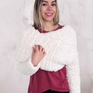 Op de afbeelding: Een witte, pluche, getailleerde trui met lange mouwen. De trui wordt gedragen over een bordeauxrode T-shirt en een zwarte jeans.