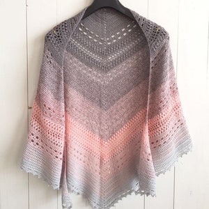May include: A handmade, open-weave shawl in a gradient of gray and pink hues. The shawl is crocheted with a chevron pattern, and has a scalloped edge. The garment is displayed on a black hanger against a white background.