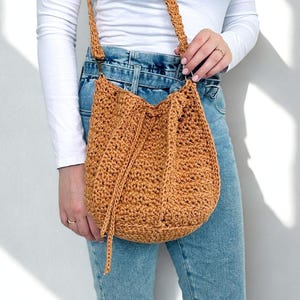 Pode incluir: Uma bolsa de ombro de crochê em um tom marrom dourado quente. A bolsa apresenta um fecho de cordão e uma alça longa. A costura texturizada cria um visual único e artesanal. A bolsa é usada no ombro.