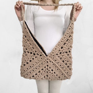Op de afbeelding: Een beige gehaakte tote bag met een V-vormige opening en een zwarte voering. De tas wordt door een persoon vastgehouden.