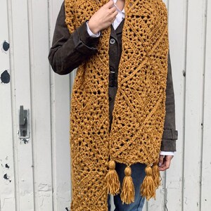 Puede incluir: Una larga bufanda de crochet de color amarillo mostaza con un patr&oacute;n geom&eacute;trico y borlas.