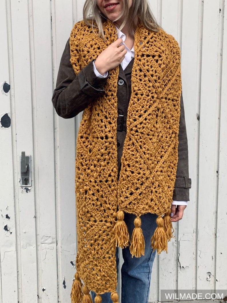 Puede incluir: Una larga bufanda de crochet de color amarillo mostaza con un patr&oacute;n geom&eacute;trico y borlas.