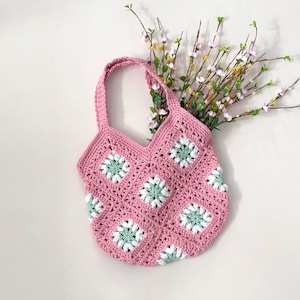 Op de afbeelding: Een roze gebreide tote bag met groene en witte bloemenaccenten. De tas heeft een brede, open bovenkant en twee lange hengsels.