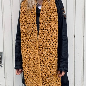 Puede incluir: Una bufanda de crochet de color amarillo mostaza con un patr&oacute;n de diamantes. La bufanda la lleva puesta una persona que lleva una chaqueta negra y unos vaqueros azules.